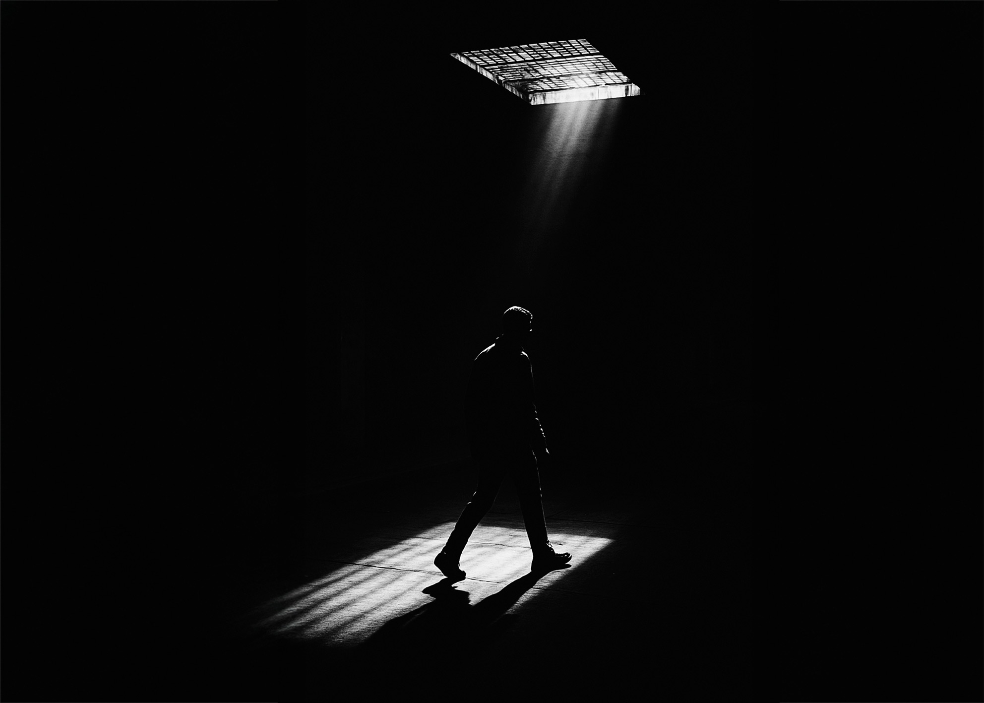 Figura maschile attraversa un cono di luce sotto un lucernario in uno spazio oscuro, la luce definisce la presenza nello spazio negativo Male figure walks through a beam of light under a skylight in a dark interior, light defines presence within negative space
