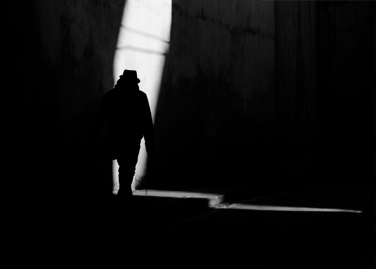 Un uomo con cappello cammina all’interno di un corridoio urbano fortemente contrastato. La luce verticale taglia lo spazio creando una tensione tra ombra e presenza, enfatizzando la solitudine e la direzione del movimento. A man wearing a hat walks through a high-contrast urban corridor. A vertical beam of light cuts through the space, creating tension between shadow and presence, emphasizing solitude and directional movement.