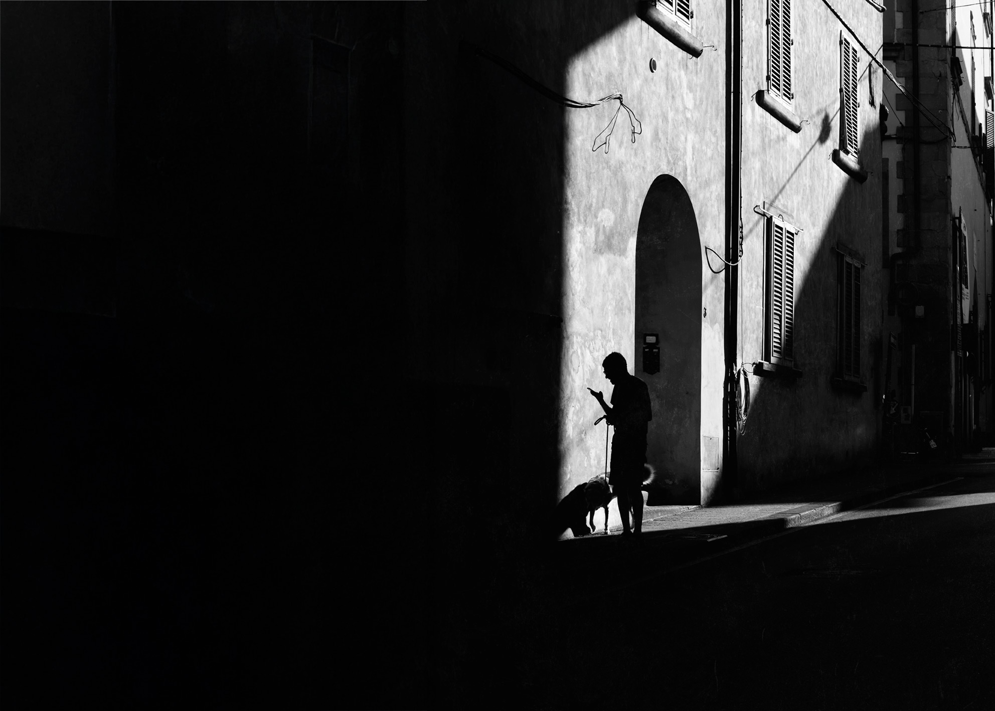 Una figura accompagna un cane lungo una diagonale architettonica netta. Il contrasto tra la superficie luminosa e la massa scura dell’edificio accentua il senso di attraversamento e silenzio metropolitano. A figure walks a dog along a strong architectural diagonal. The contrast between the bright surface and the dark mass of the building enhances the feeling of crossing and metropolitan silence.