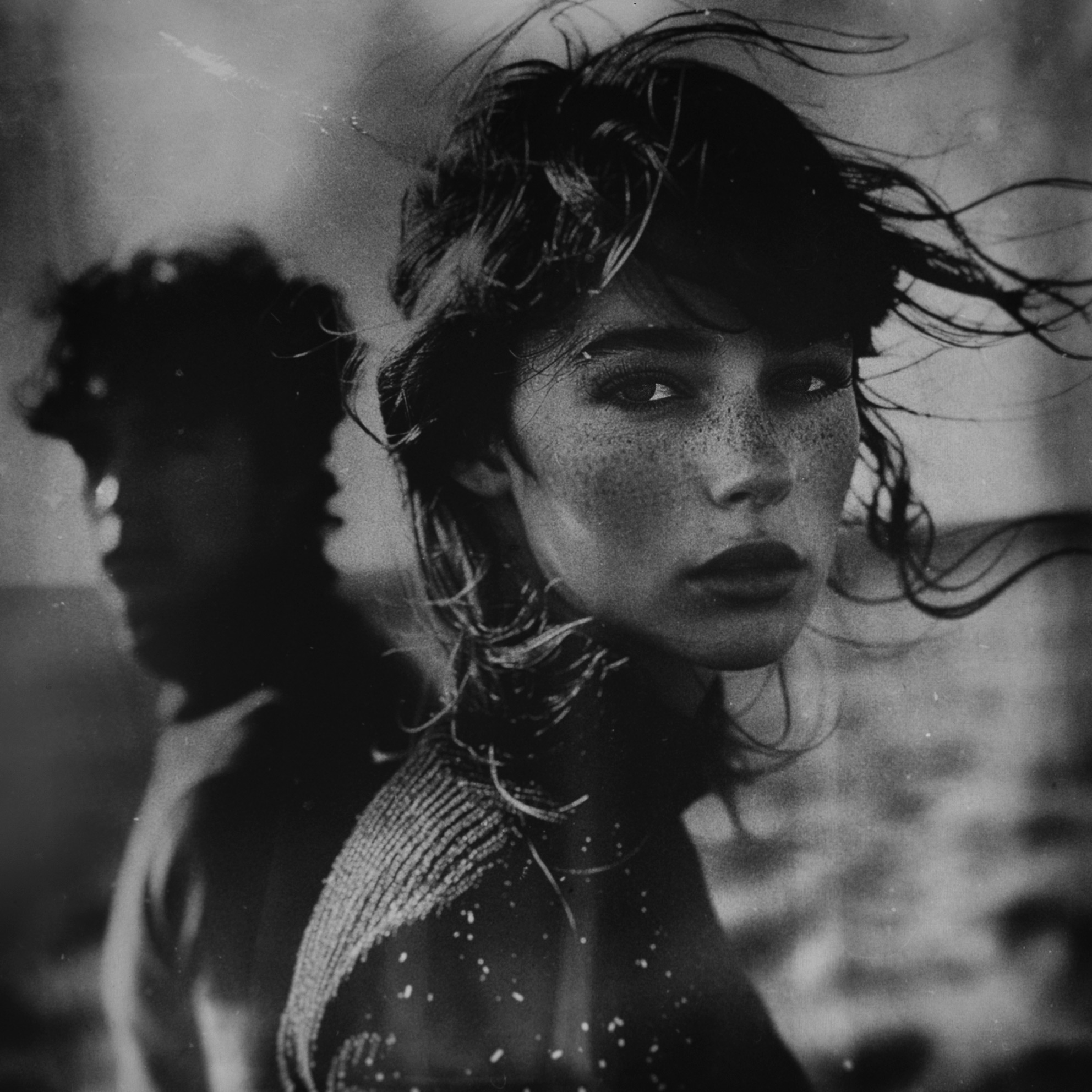 Ritratto in bianco e nero di una ragazza con lentiggini in primo piano con una figura sfocata sullo sfondo in ambiente naturale Black and white portrait of a freckled girl in foreground with a blurred figure in the background in a natural environment