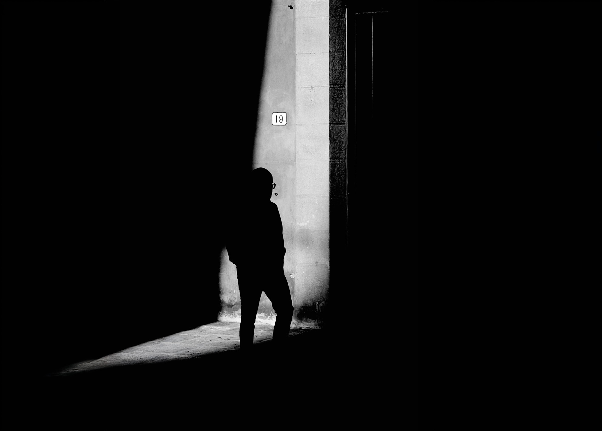 Una figura attraversa uno spazio architettonico segnato dal numero 19. La verticalità delle linee e l’ombra dominante costruiscono una scena di sospensione e misura, dove il soggetto diventa punto di riferimento visivo. A figure crosses an architectural space marked by the number 19. The vertical lines and dominant shadow build a suspended and measured scene, where the subject becomes a visual anchor.