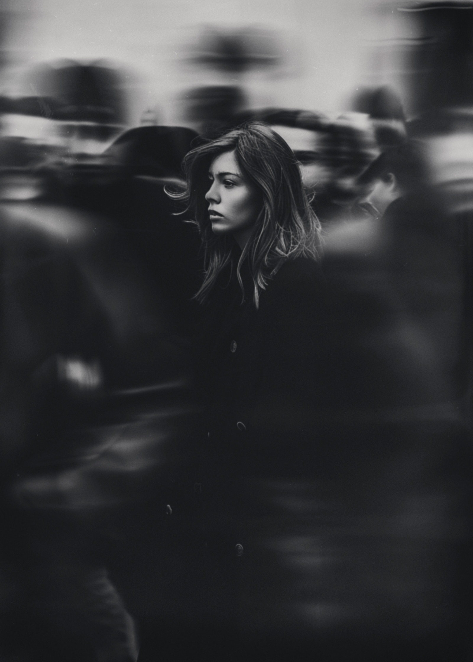 Ritratto in bianco e nero di una donna ferma mentre la folla intorno a lei si muove in modo sfocato, contrasto tra immobilità e movimento che suggerisce isolamento e concentrazione interiore, a black and white portrait of a woman standing still as a blurred crowd moves around her, a contrast between stillness and motion evoking isolation and inner focus.
