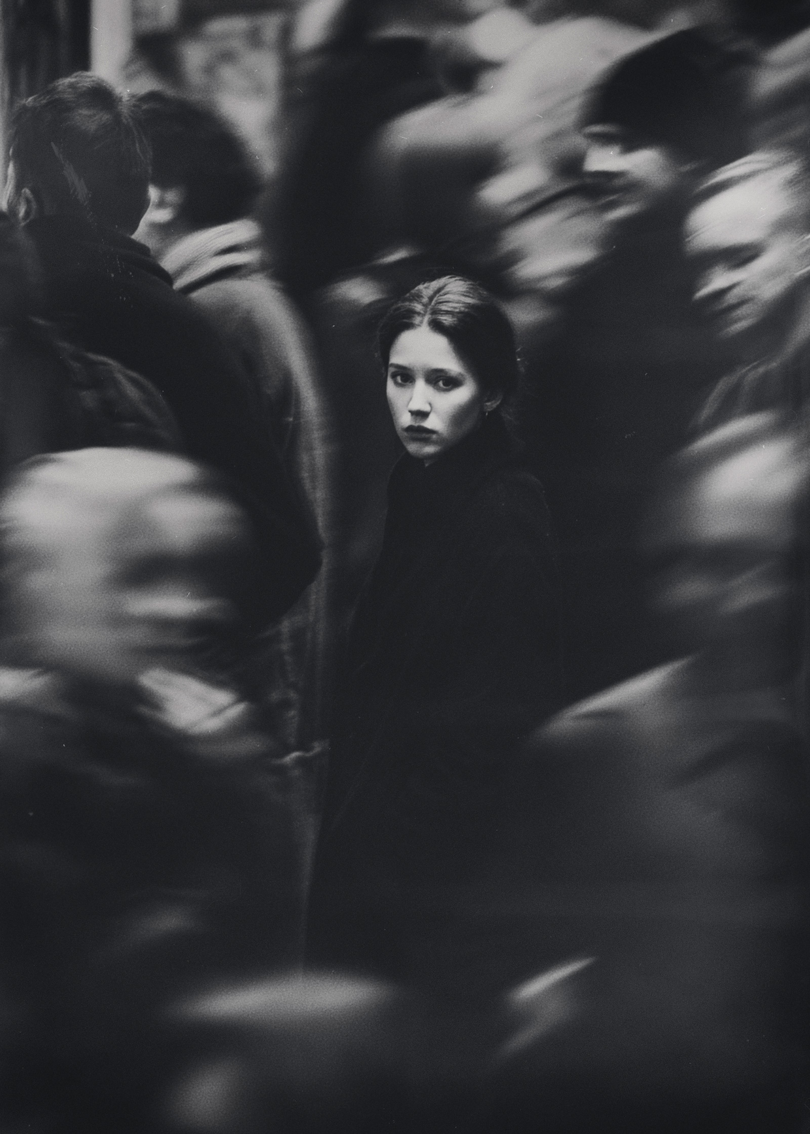 Figura femminile al centro di una folla in movimento, lo sguardo diretto verso l’obiettivo mentre il contesto circostante appare mosso e indefinito, creando una tensione visiva tra presenza e anonimato, a female figure centered within a moving crowd, looking directly at the camera while the surroundings blur, creating visual tension between presence and anonymity.